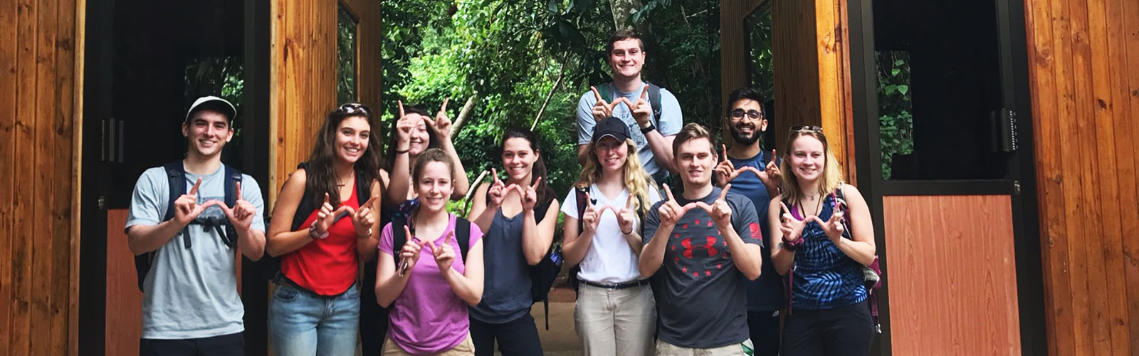 Ten students standing in a walkway between two wooden buildings, holding up the letter "W" with their fingers.